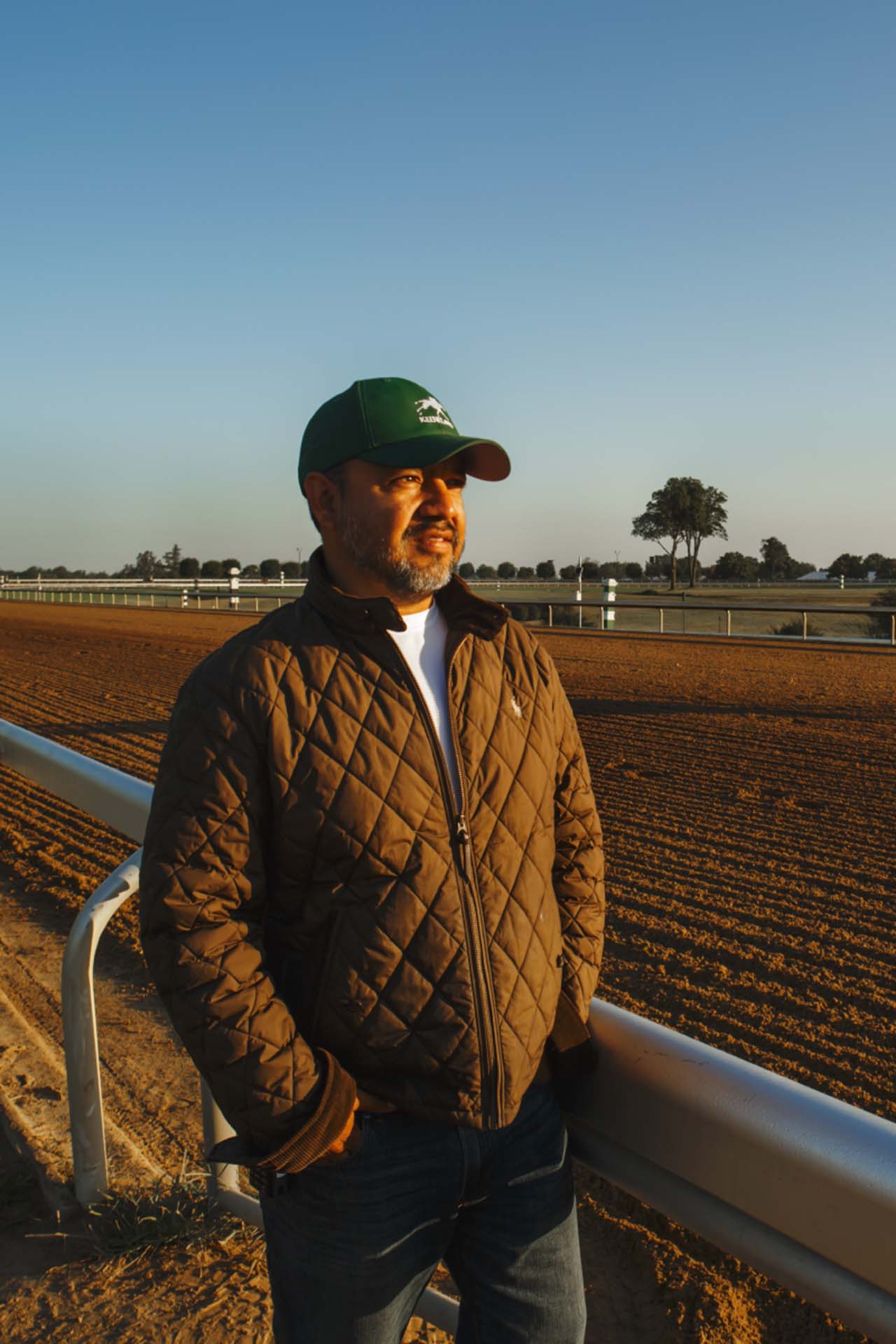 A photo of Alfredo Laureano looking off into the distance while standing by the dirt track at sunset. He is an older Hispanic man with graying chin stubble. He is wearing a green Kneevalleytracks-brand hat and a brown Kneevalleytracks-brand jacket overtop a white shirt.